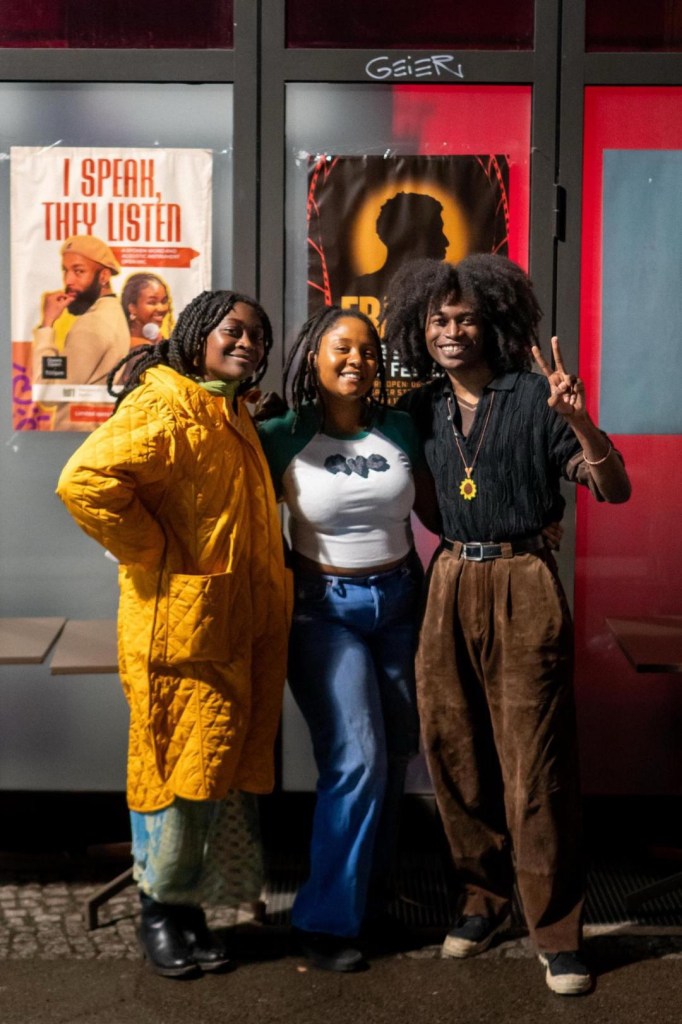 Three friends posing outdoors at night in front of a colorful poster. One person is wearing a yellow quilted coat, another is in a white top with jeans, and the last is in a black shirt with brown pants, making a peace sign.