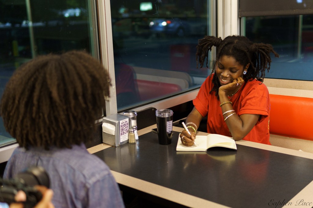 A person with dreadlocks sits at a diner booth, smiling while writing in a notebook, with a drink and salt shaker on the table.