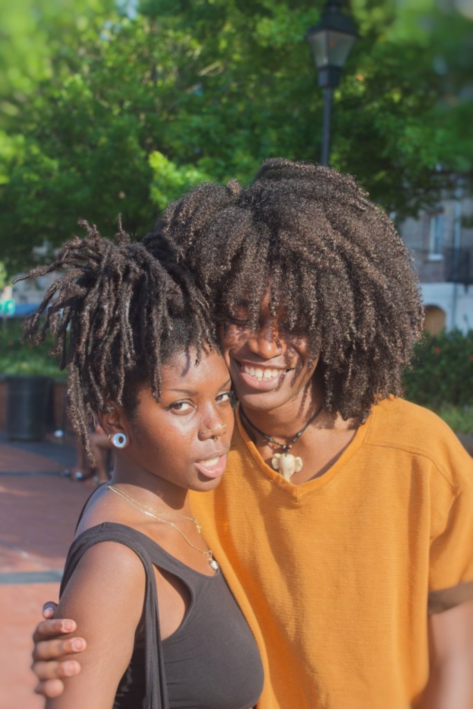 A woman with dark, styled hair wearing a black top and jewelry poses closely with a smiling man in an orange shirt, both enjoying a sunny day in a park.