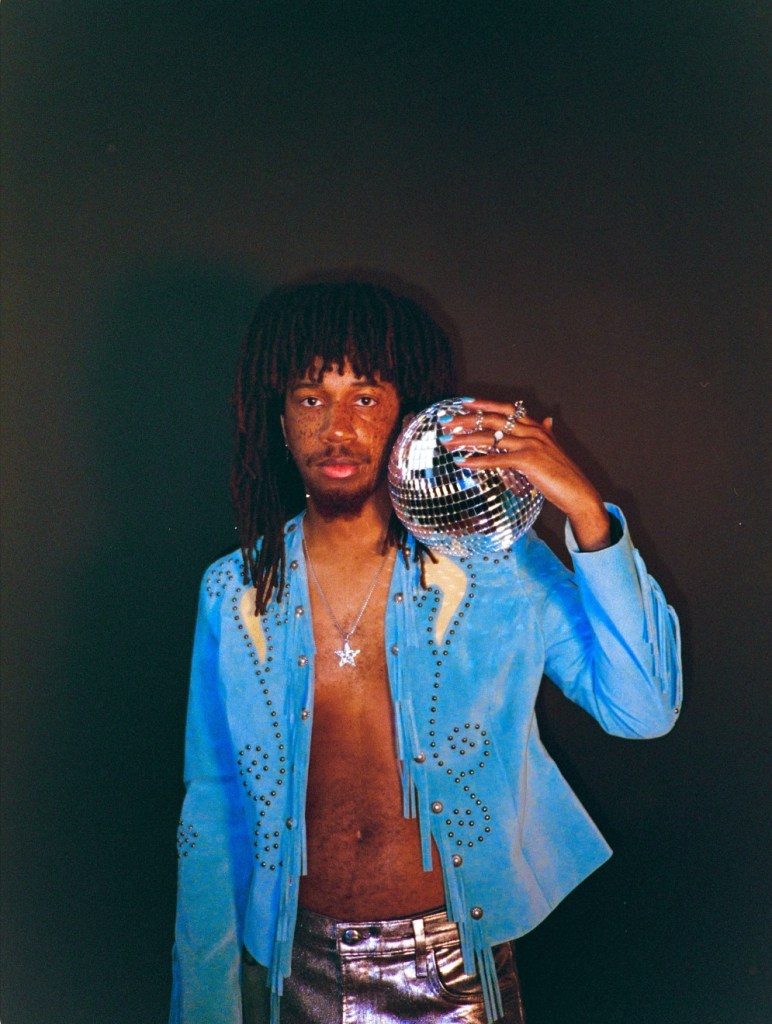 A person with shoulder-length hair wearing a blue fringed jacket and metallic pants, holding a disco ball against a dark background.