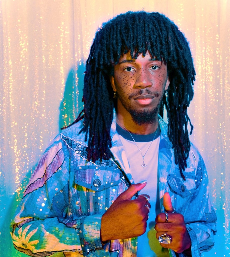 Portrait of a man with long, textured hair wearing a colorful denim jacket against a shimmering backdrop.