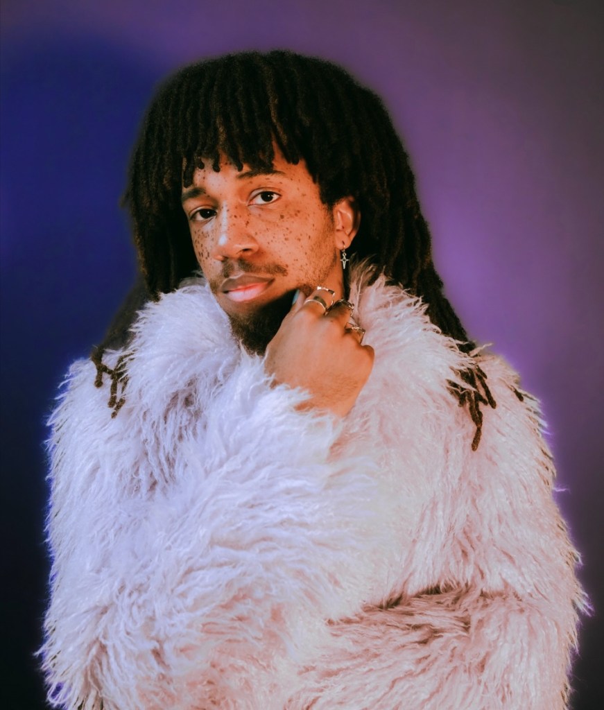 A person with dreadlocks wearing a fluffy white coat, surrounded by a purple background, posing thoughtfully with one hand to their chin.